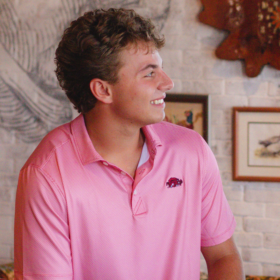 A close-up of a red polo shirt with a subtle geometric pattern, featuring a small embroidered red and black Arkansas Razorbacks Slobbering Hog logo on the chest.