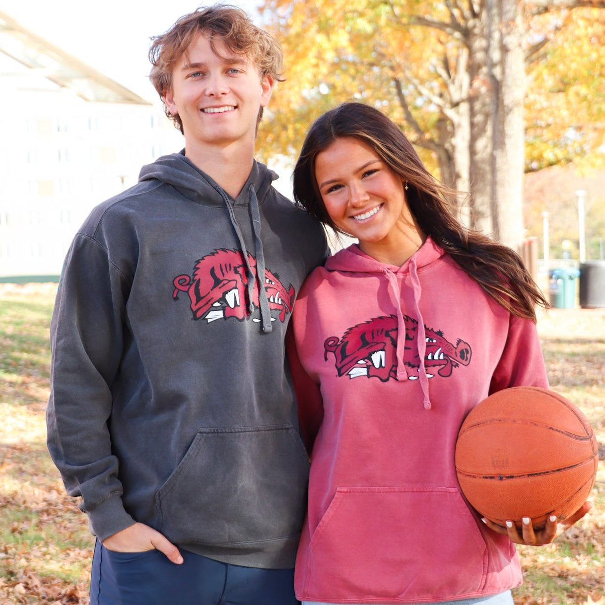 Two people wearing Arkansas Razorback Houndstooth hoodies with the iconic slobbering hog logo, one holding a basketball, standing outside Bud Walton Arena with trees in the background.