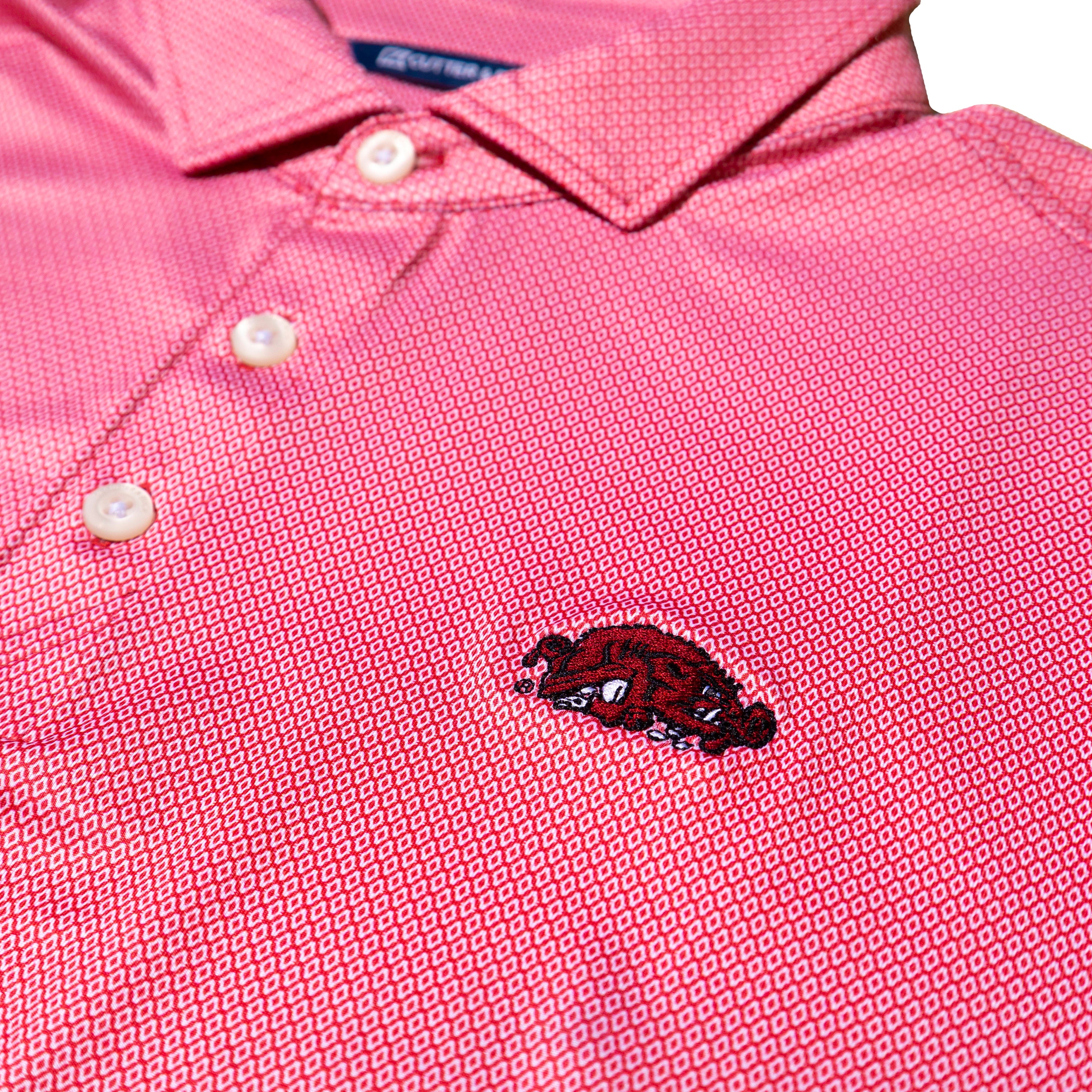 A close-up of a red polo shirt with a subtle geometric pattern, featuring a small embroidered red and black Arkansas Razorbacks Slobbering Hog logo on the chest.