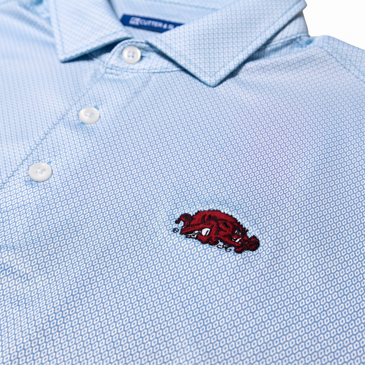 Close-up of a light blue patterned polo shirt with a red embroidered Arkansas Razorbacks logo on the chest.