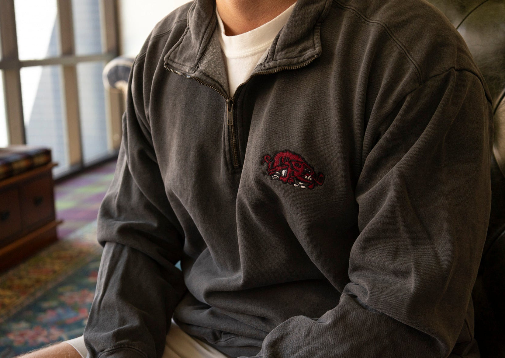 A person seated indoors wearing a dark charcoal quarter-zip sweatshirt with a red embroidered Arkansas Razorbacks basketball logo on the left chest.
