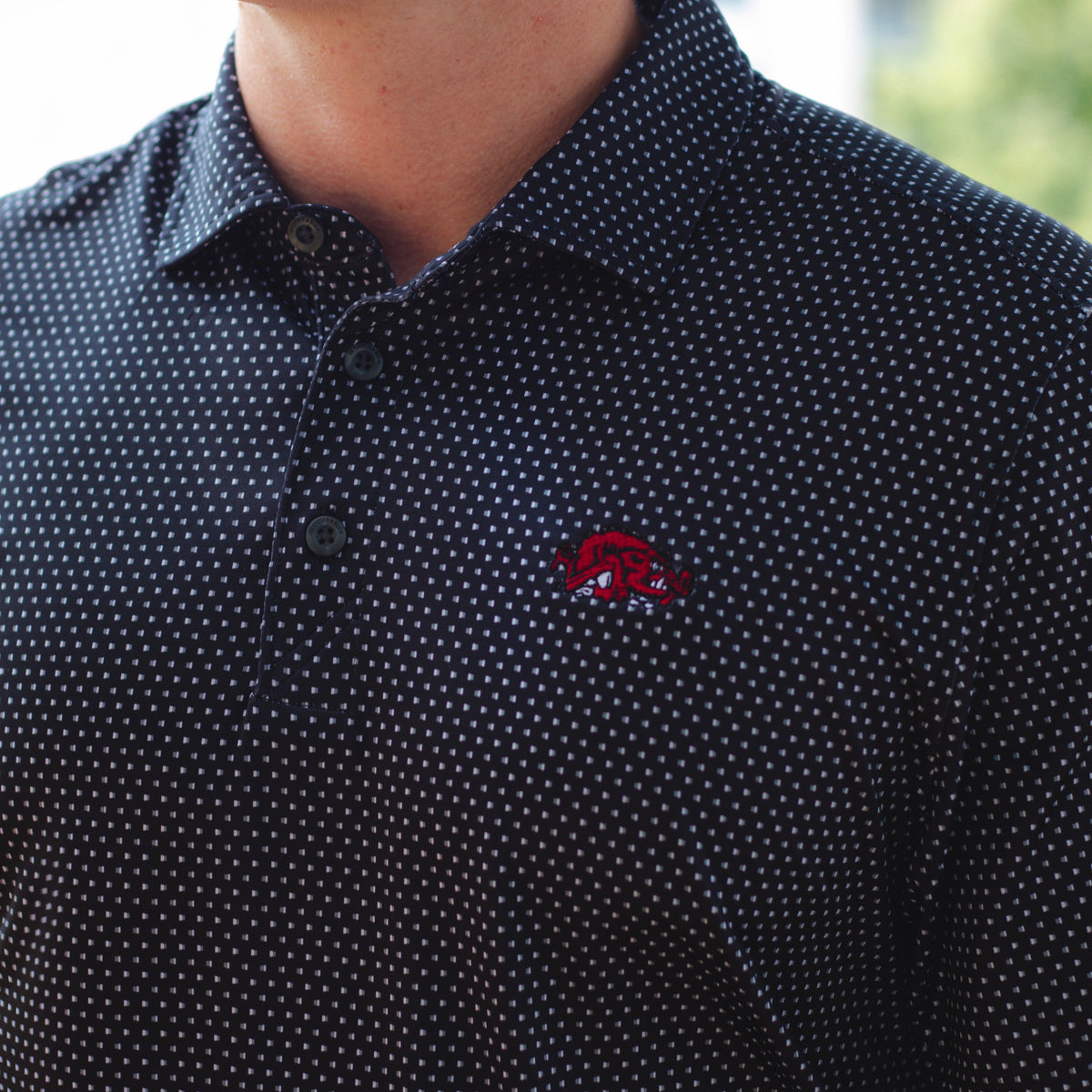 A close-up of a man wearing a black short-sleeve polo shirt with a small white micro-dot pattern and a red embroidered Razorback logo on the left chest.