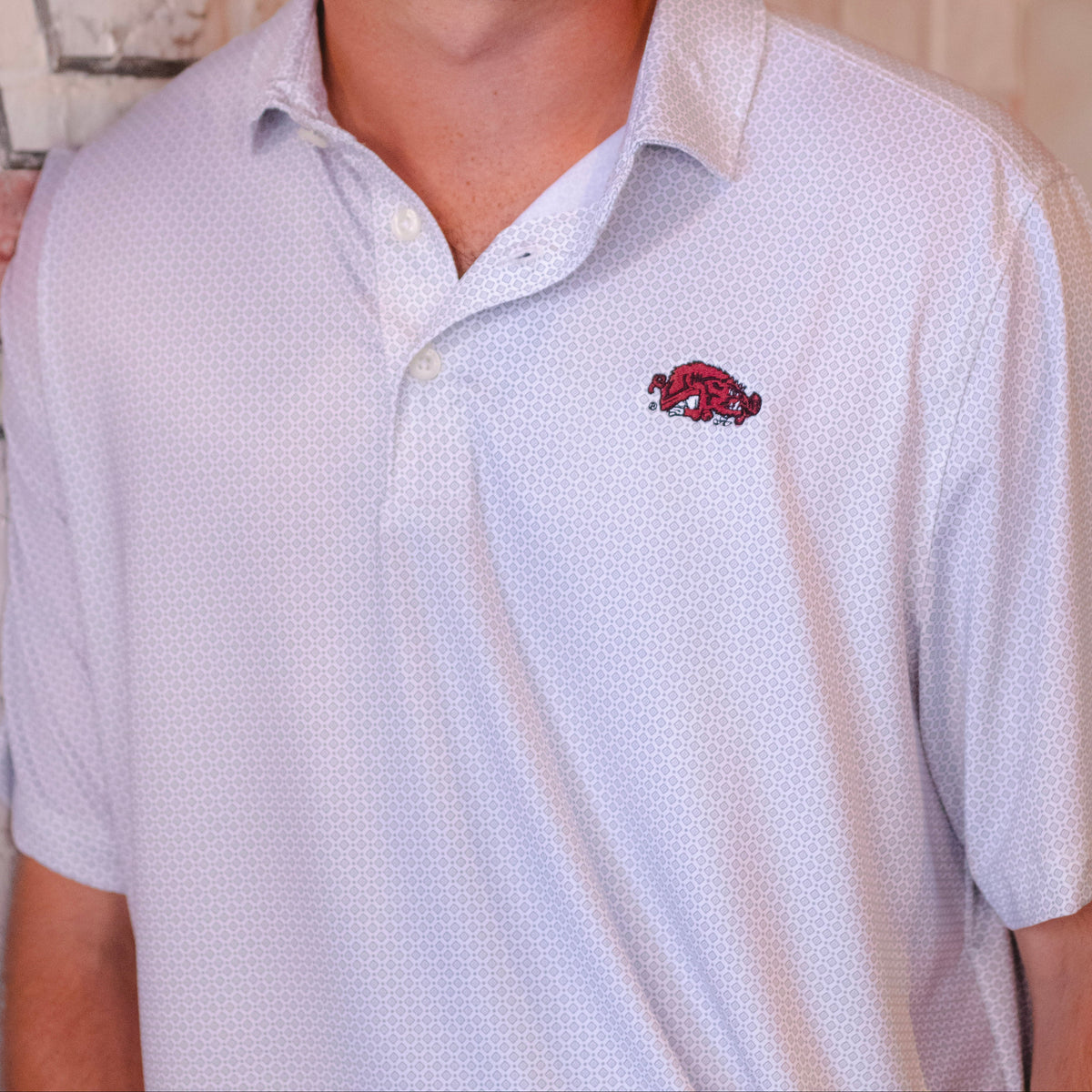 A smiling young man with dark hair wearing a white short-sleeve polo shirt with a small red and black Arkansas Razorbacks logo on the left chest, standing against a white brick wall.