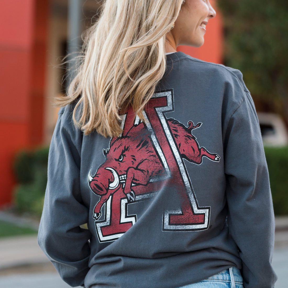 A dark charcoal gray long-sleeve t-shirt featuring a large red, white, and black graphic of the University of Arkansas Razorbacks 'A' with a running razorback on the front, displayed against a light off-white background.