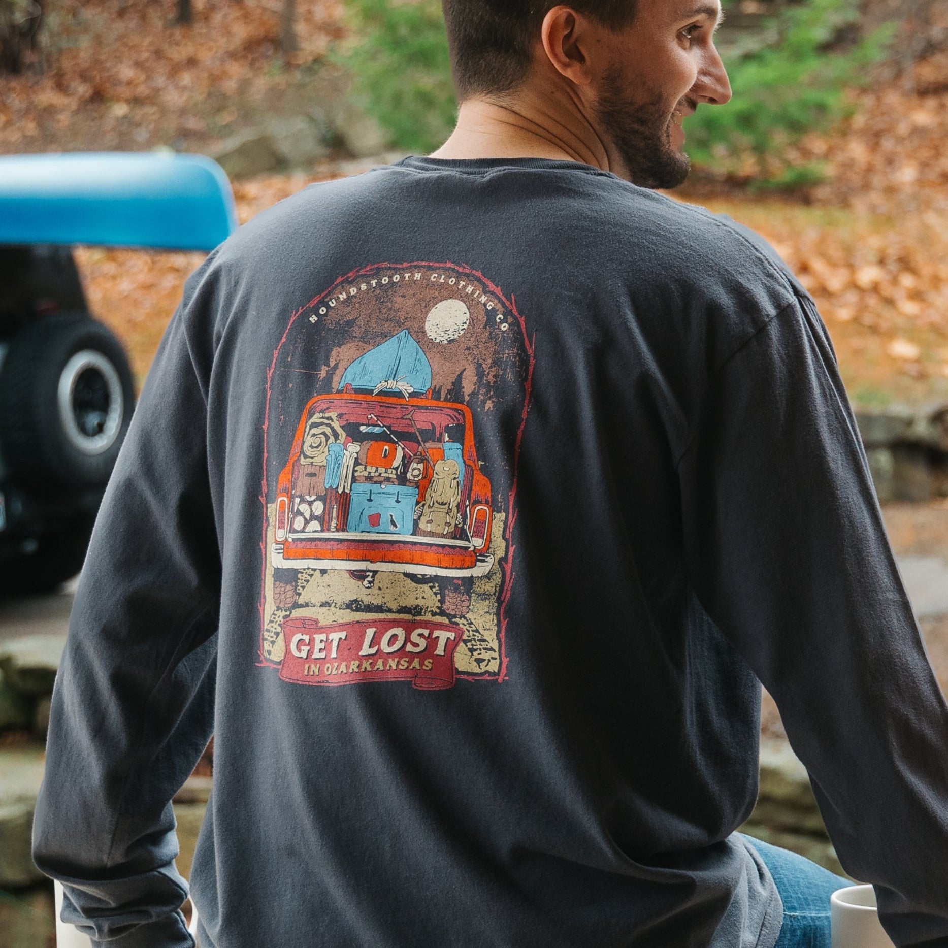 Vintage jeep packed full of camping gear, hiking backpack, cooler, firewood, tents, blankets, a canoe on top, underneath a full moon. Get Lost in Ozarkansas displayed underneath.