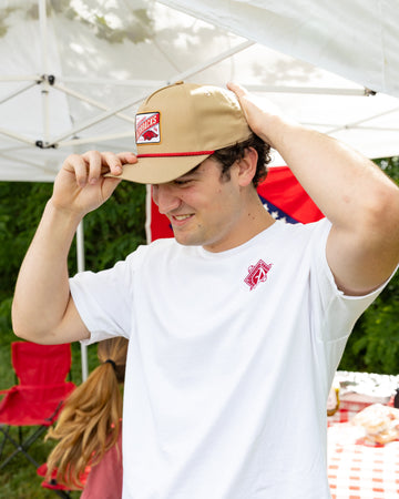 Person wearing a beige cap with a red logo and white t-shirt, standing outdoors under a white canopy.