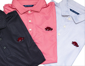 Three folded short-sleeve polo shirts shown on a white background: a dark black shirt with small white dots, a red shirt, and a light grey shirt with a subtle white dot pattern. Each shirt has a small embroidered red Razorback basktball logo on the chest.