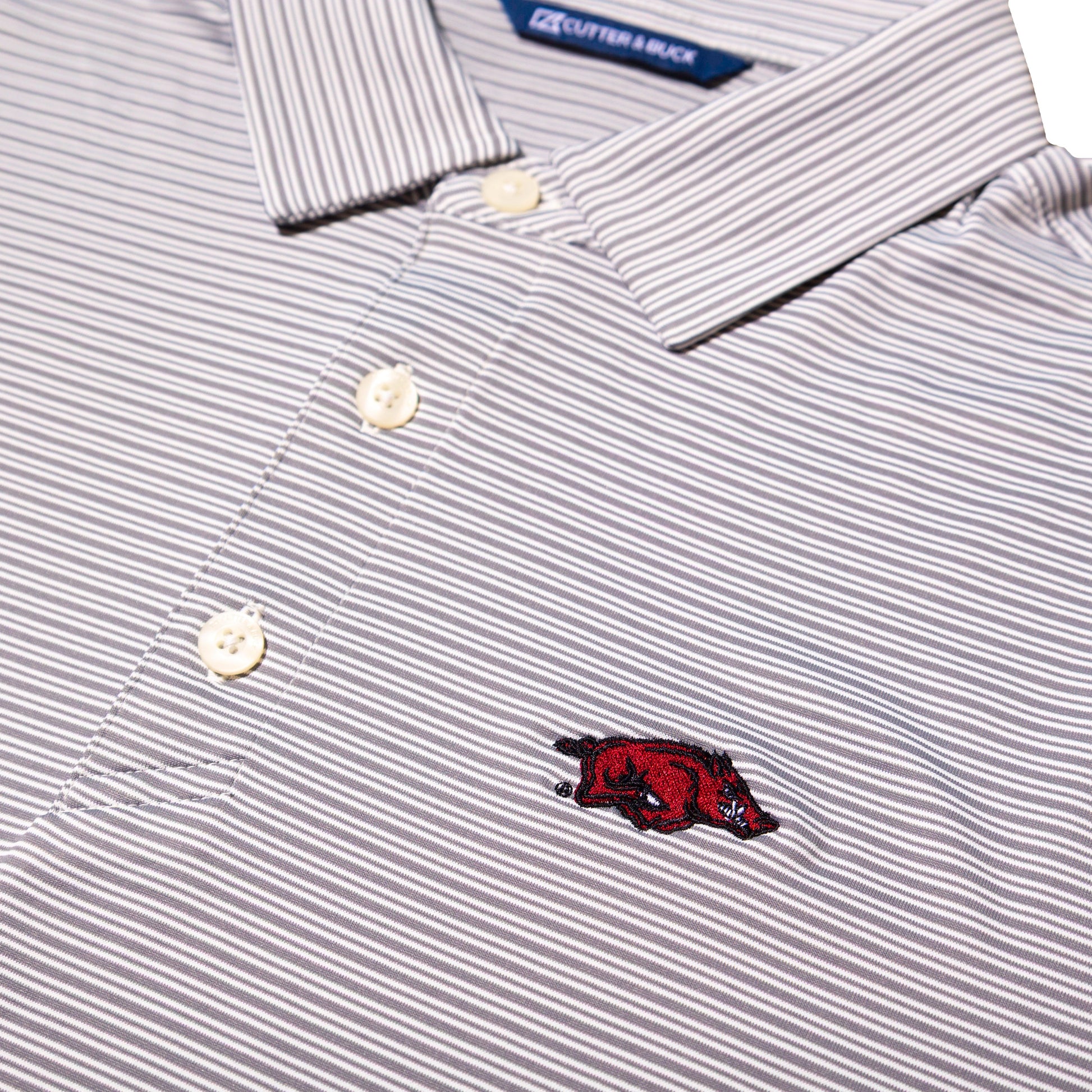 Close-up of a striped polo shirt with thin gray and white vertical pinstripes, cream buttons, and a small embroidered red razorback logo on the chest.