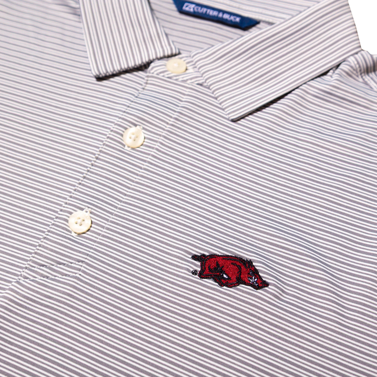 Close-up of a striped polo shirt with thin gray and white vertical pinstripes, cream buttons, and a small embroidered red razorback logo on the chest.