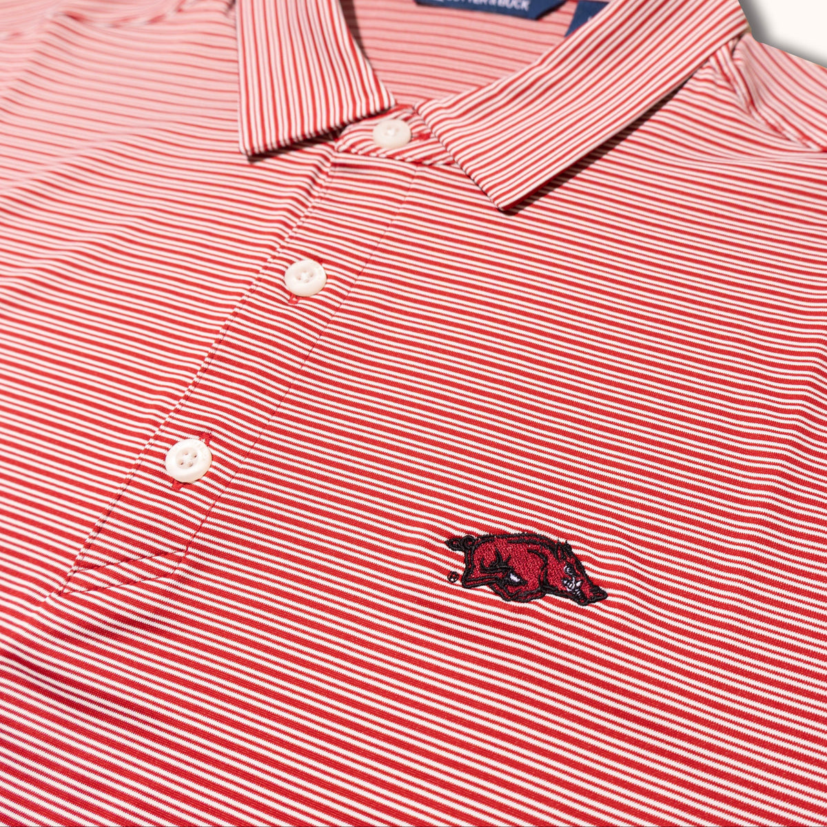 A close-up of a red and white striped polo shirt with a small embroidered red Razorback logo on the chest, set against a clean off-white background.