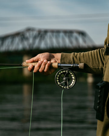 Person holding a fly fishing rod with the Cotter, AR bridge blurred and the water of the White River in the background