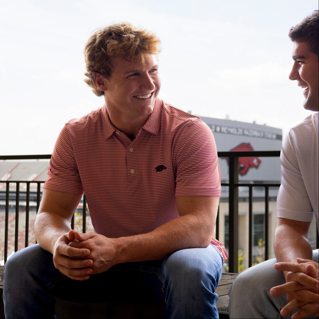 Two men sitting and smiling outdoors, wearing matching polo shirts with a small Arkansas Razorbacks logo on the chest.